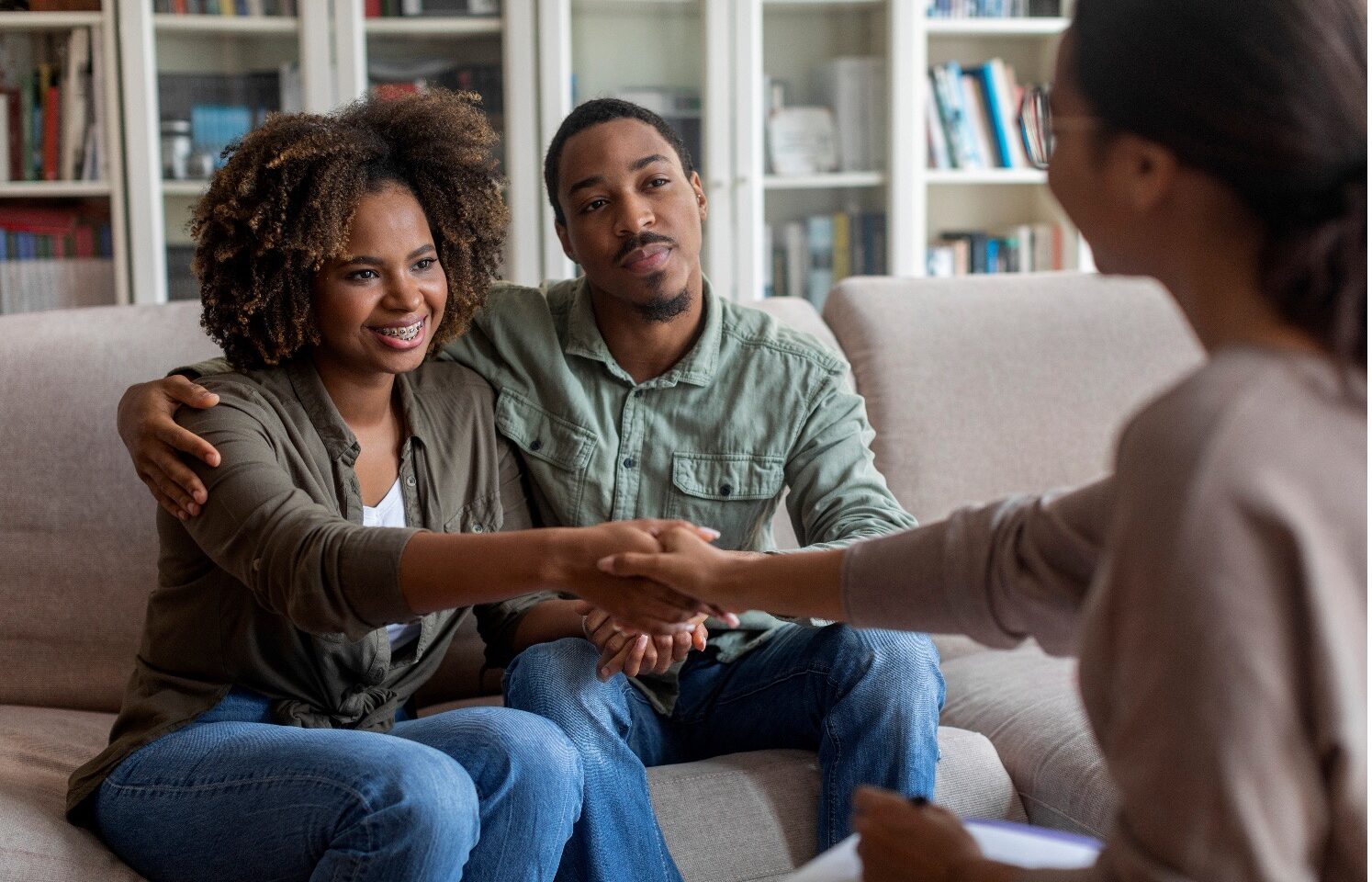 Couple sits on a beige sofa in a living room, smiling and shaking hands with a counselor across from them in a therapy session.