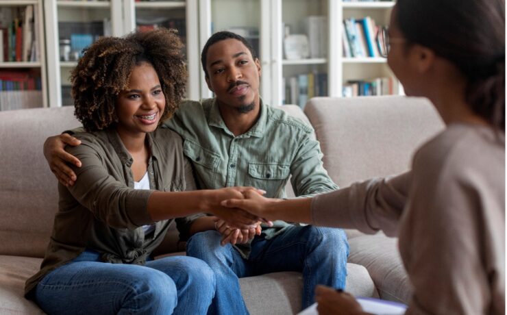 Couple sits on a beige sofa in a living room, smiling and shaking hands with a counselor across from them in a therapy session.