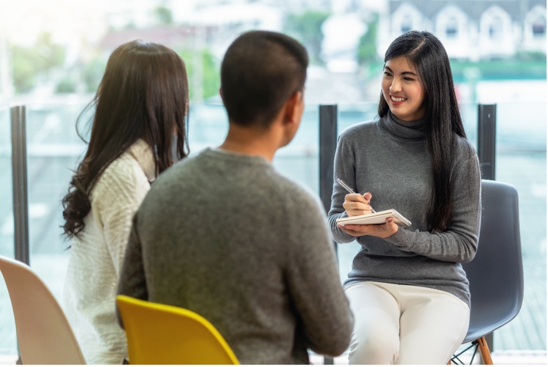 Three adults in a bright office; a smiling woman in a gray turtleneck writes in a notebook as two people sit opposite her for a meeting or counseling session.