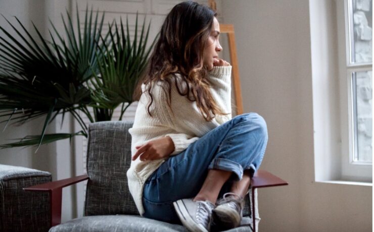 Woman sitting cross-legged on a gray chair by a window, resting her chin on her hand in a thoughtful pose.