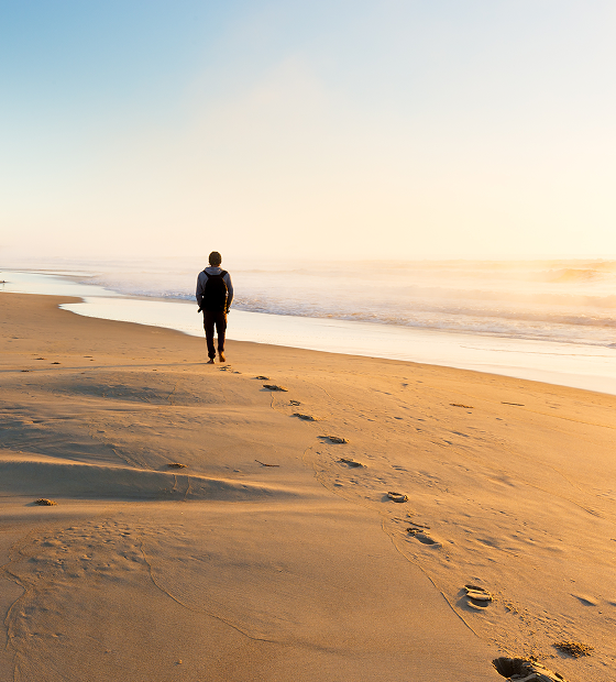 Person with a backpack walks along a sandy beach toward the ocean at sunset, leaving footprints in the wet sand.