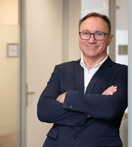 Professional portrait of a man in a navy suit and glasses, arms crossed in an office setting.