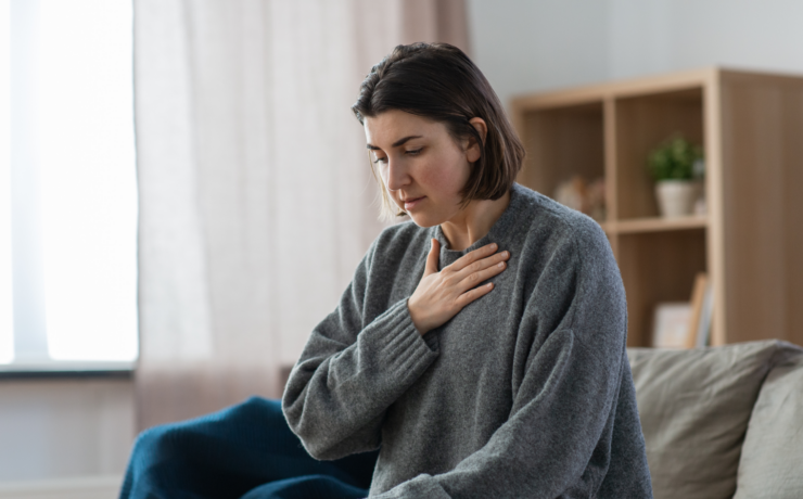Woman sitting on a sofa, hand pressed to her chest in quiet discomfort at home.