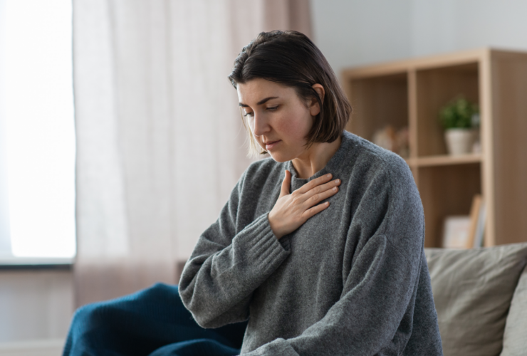 Woman sitting on a sofa, hand pressed to her chest in quiet discomfort at home.