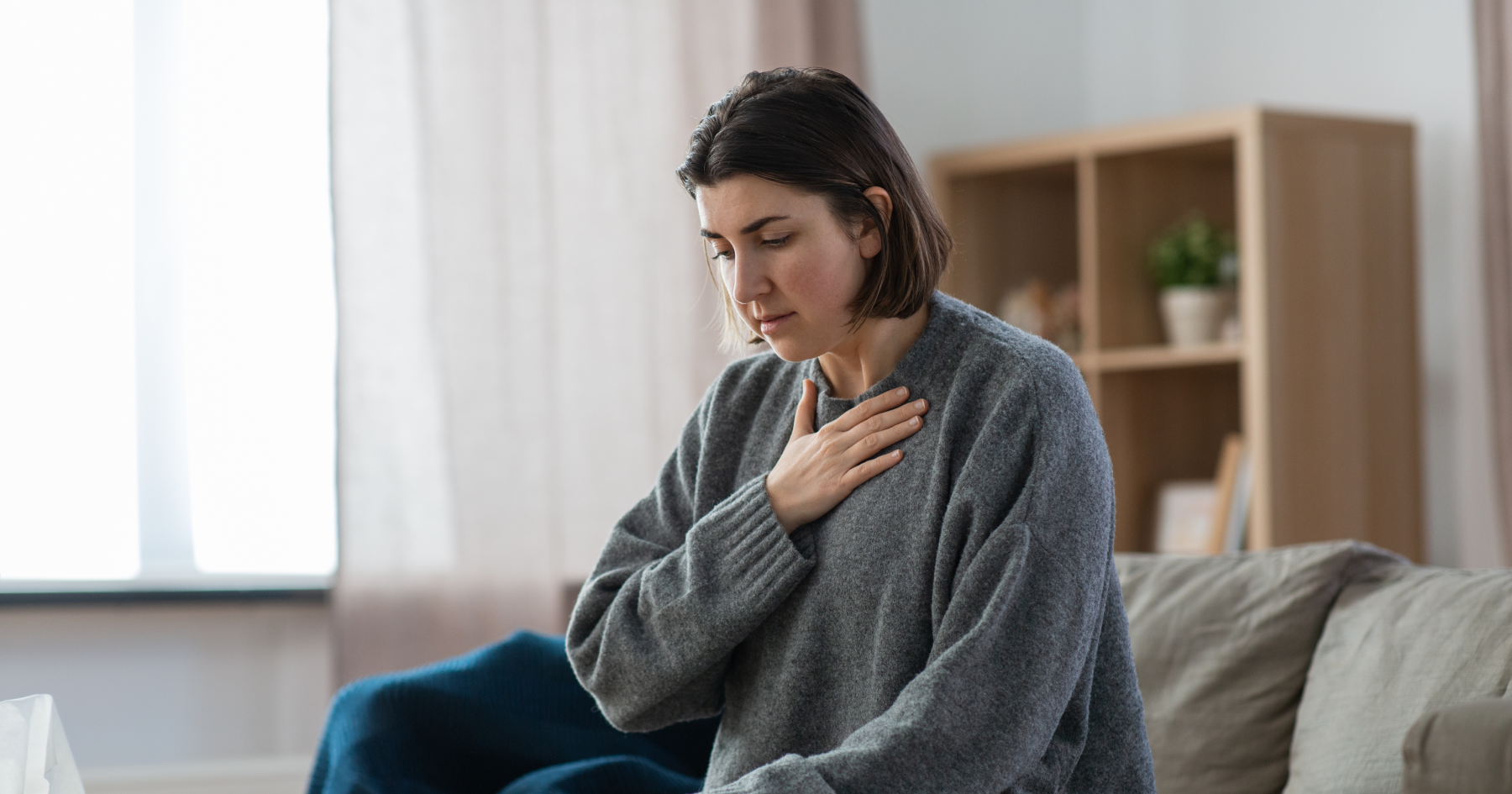 Woman sitting on a sofa, hand pressed to her chest in quiet discomfort at home.