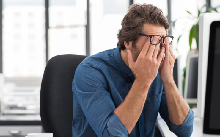 Man in a blue shirt sits at a desk, rubbing his eyes in frustration at the computer.