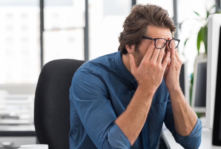 Man in a blue shirt sits at a desk, rubbing his eyes in frustration at the computer.