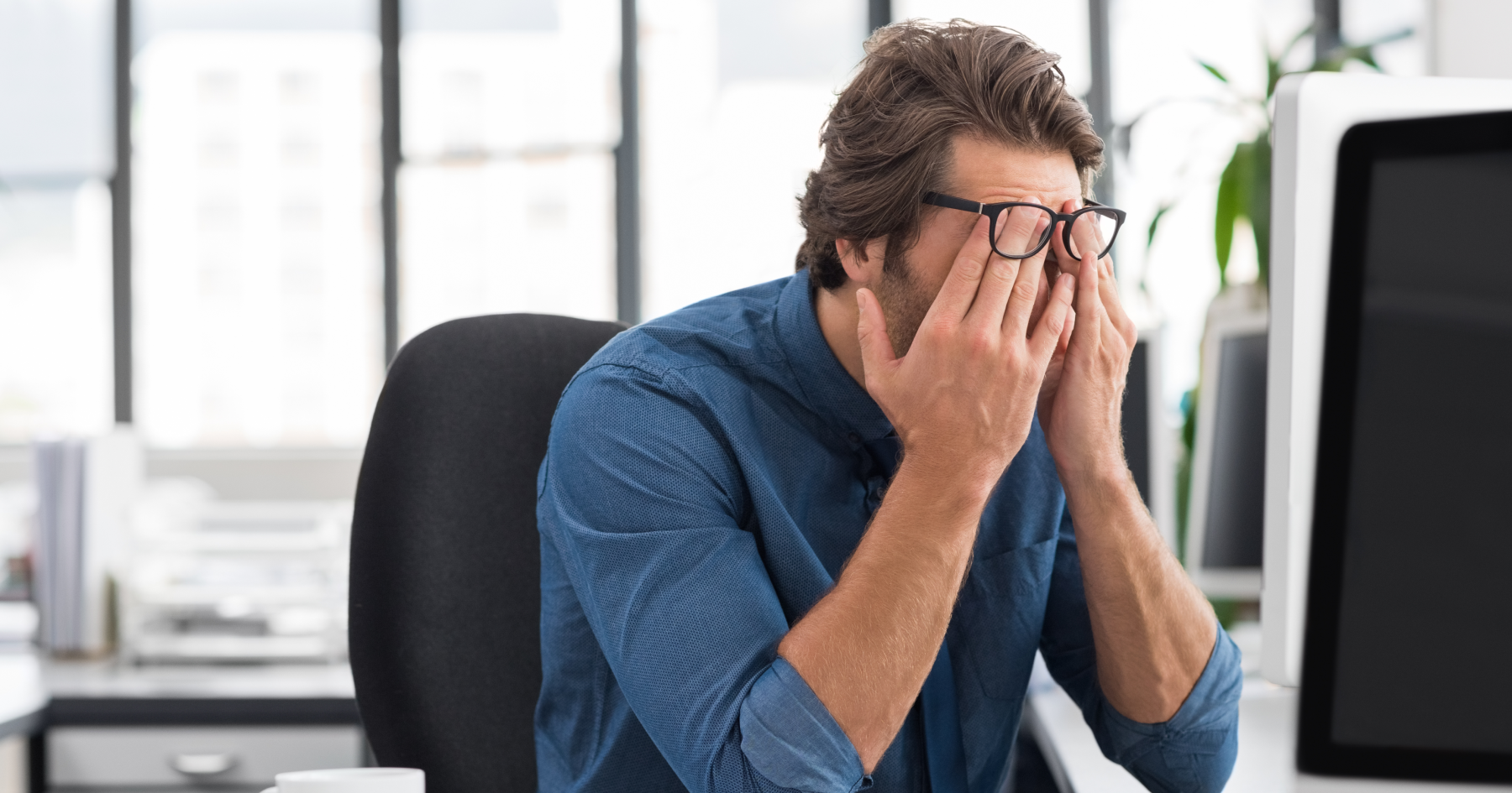 Man in a blue shirt sits at a desk, rubbing his eyes in frustration at the computer.