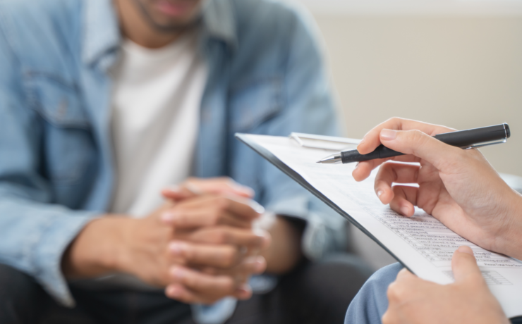 Clinician’s hand with a pen poised over a clipboard during a counseling session; client sits across, hands clasped.