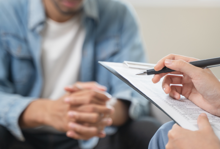 Clinician’s hand with a pen poised over a clipboard during a counseling session; client sits across, hands clasped.