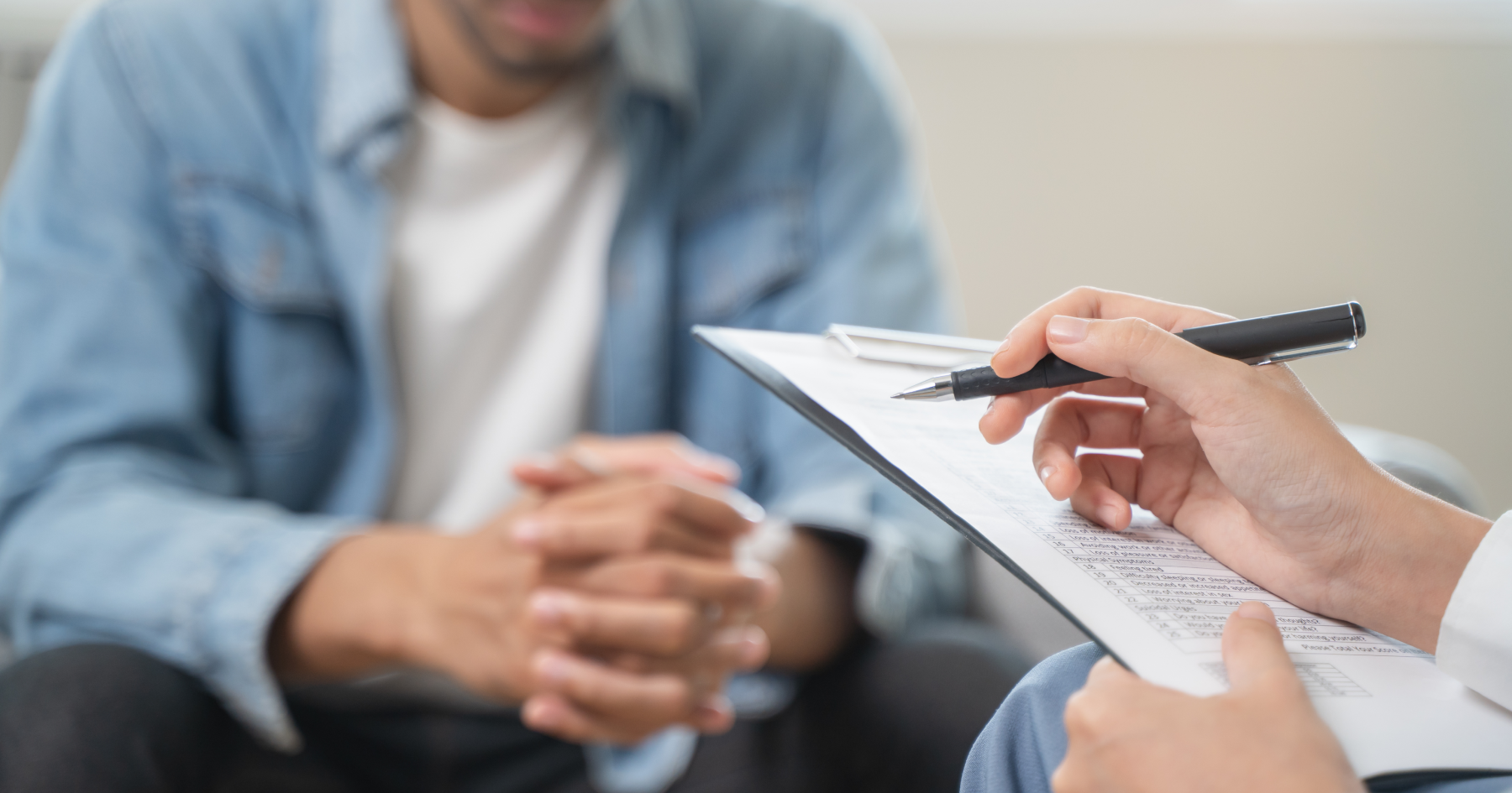 Clinician’s hand with a pen poised over a clipboard during a counseling session; client sits across, hands clasped.